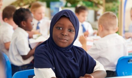 A child wearing a hijab, looking to camera whilst the class continues around them